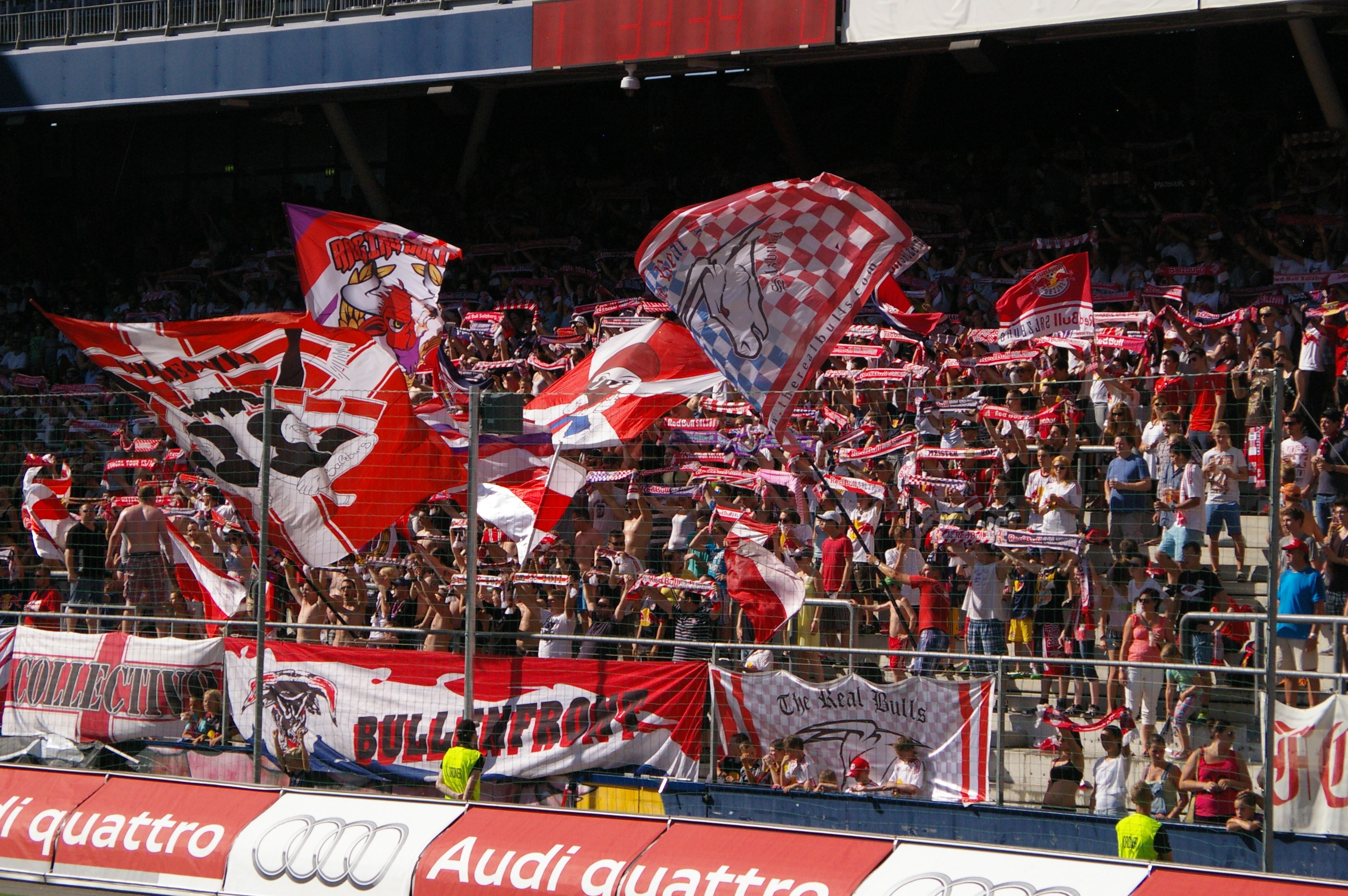 Eine große Menschenmenge in einem Stadion, einige halten Fahnen und Banner mit Text, andere stehen auf dem Boden, umgeben von einem Metallzaun, einem Schild, einem Anzeigebildschirm und einer Decke mit Deckenleuchten.