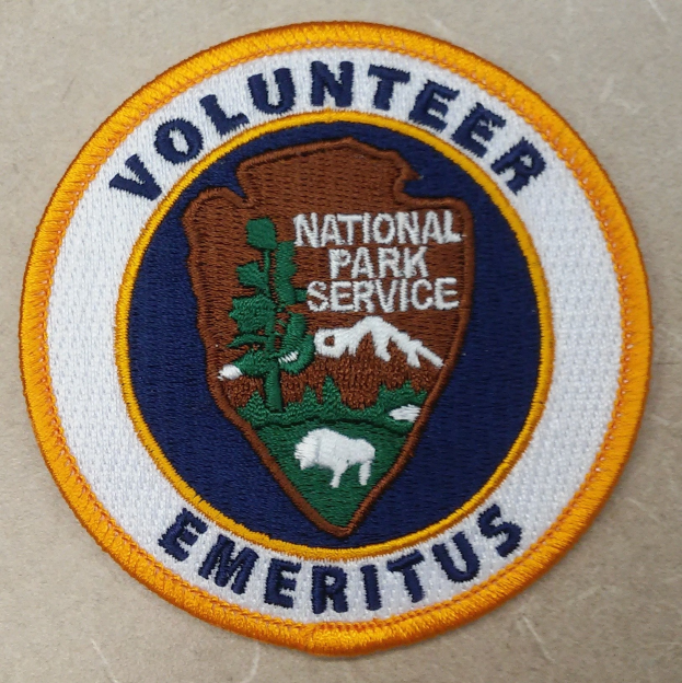 Runder Aufnäher mit blauem Rand und weißem Hintergrund, der mittig den schwarzen Text "Volunteer National Park Service Emeritus" in einem gelben Kreis zeigt.