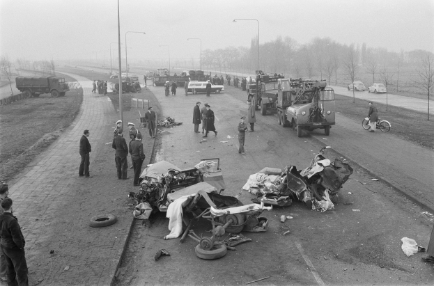 Schwarz-weißes Bild eines Autounfalls am Straßenrand mit mehreren Fahrzeugen und einer Gruppe von Menschen drumherum, Lichtmasten, Bäume und Himmel im Hintergrund.