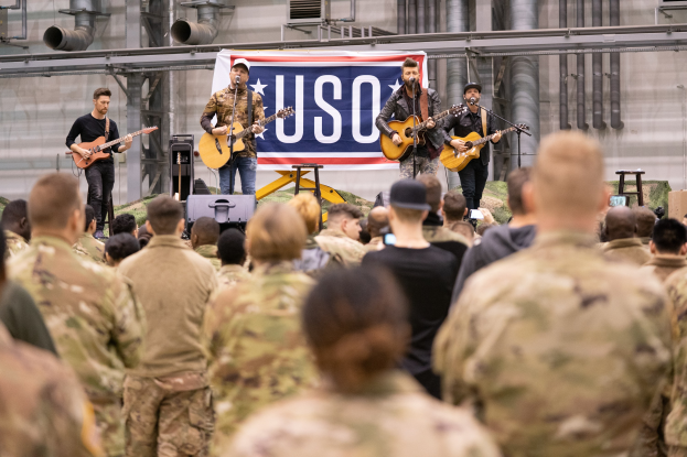 Eine Gruppe von Soldaten performs mit Gitarren und Mikrofonen für ein Publikum, mit einem Banner und Pfeifen im Hintergrund.