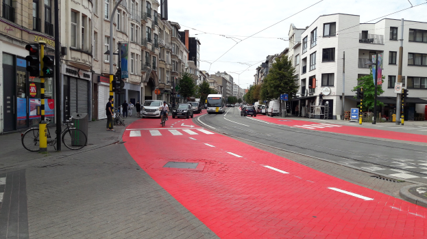 Eine Stadtstraße mit einer roten Fahrradspur, Fahrzeugen, Fußgängern, Gebäuden, Bäumen und einem klaren blauen Himmel.