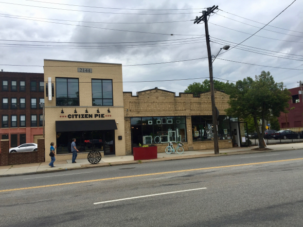Eine belebte Stadtstraße mit Fußgängern, parkenden Fahrrädern, Strommasten, Bäumen und Gebäuden, mit dem Restaurant Citizen Pie an der Ecke unter einem bewölkten Himmel.