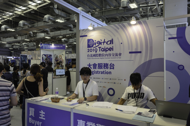 Eine Gruppe von Menschen an einem Messestand mit zwei sitzenden Personen, einem Tisch mit Gegenständen und Hintergrundplakaten mit der Aufschrift "Digital 2019 Taipei International Digital Content Summit & Fair."