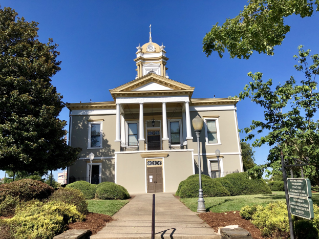 Großes Backsteingebäude mit einem Turm, umgeben von Fenstern, Türen, Säulen und einem Dach, das als das alte Rathaus bezeichnet wird, das kürzlich wiedereröffnet wurde, mit Straßenmöbeln, Grünflächen und einem bewölkten Himmel.