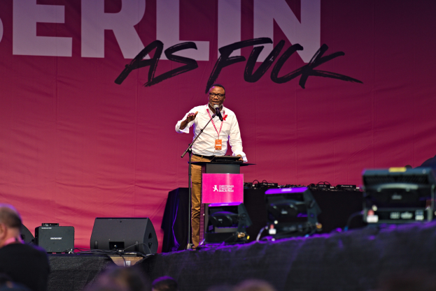 Mann in weißem Hemd und braunen Hosen, der in ein Mikrofon spricht, vor einem pinken 'Berlin as Fuck'-Plakat