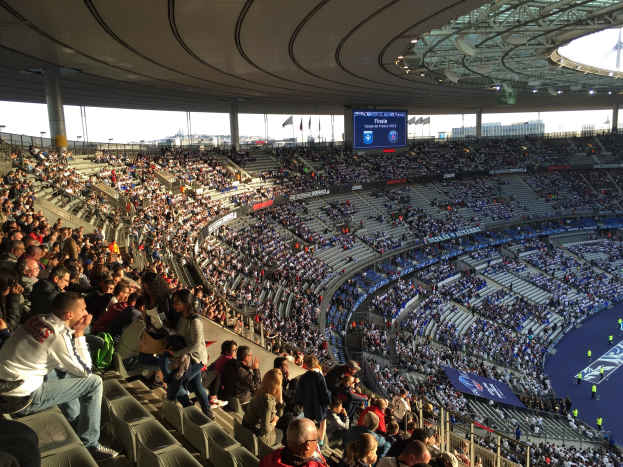 Große Menschenmenge in einem Stadion bei einem Fußballspiel mit einer Bühne rechts, Fahnen, Stangen, einem Bildschirm und der Allianz Arena in München, Deutschland im Hintergrund.