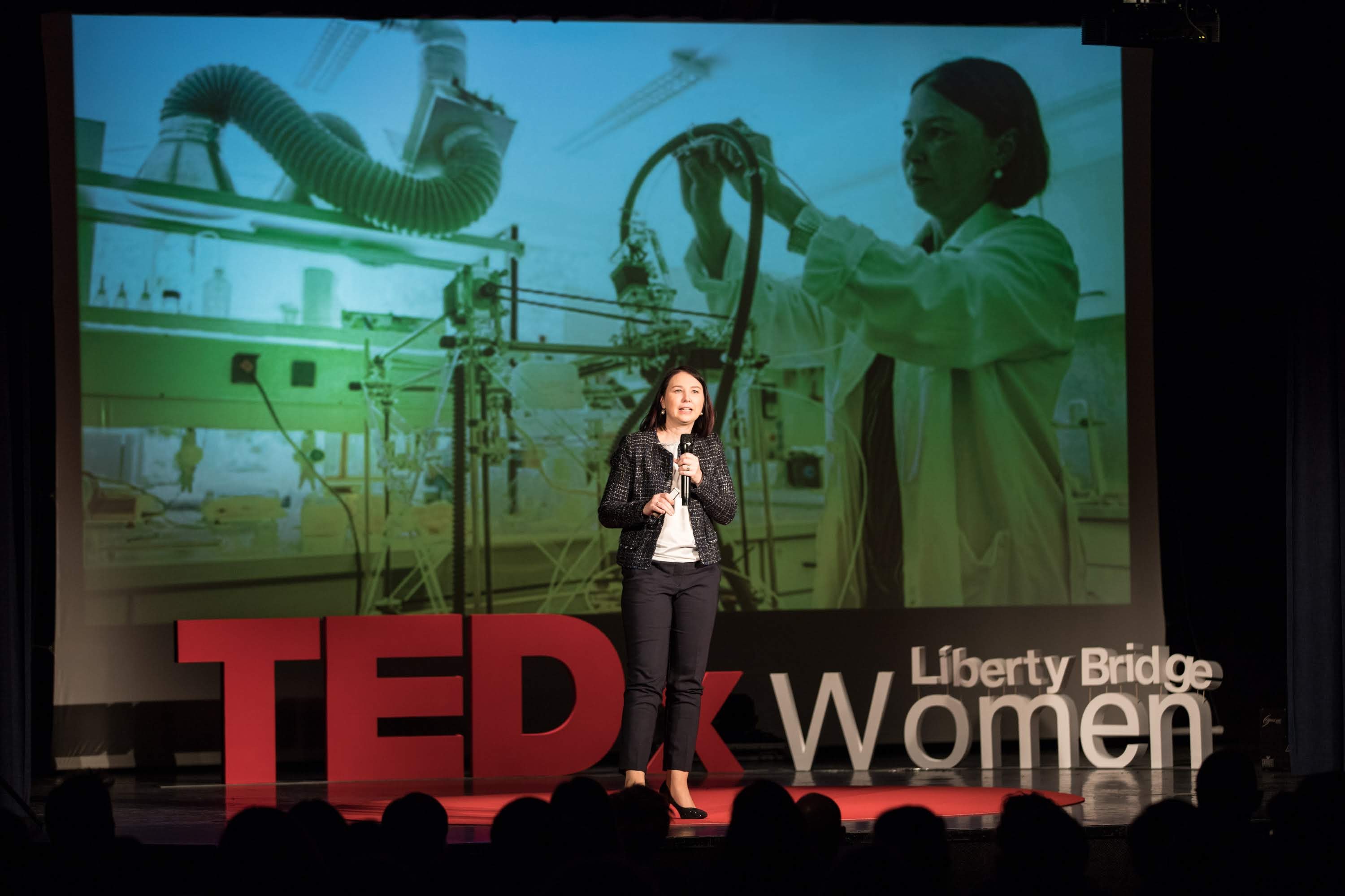 Frau auf einer Bühne mit einem Mikrofon, hinter ihr ein Display mit einem Bild einer Frau und Text zum TEDxWomen-Event.