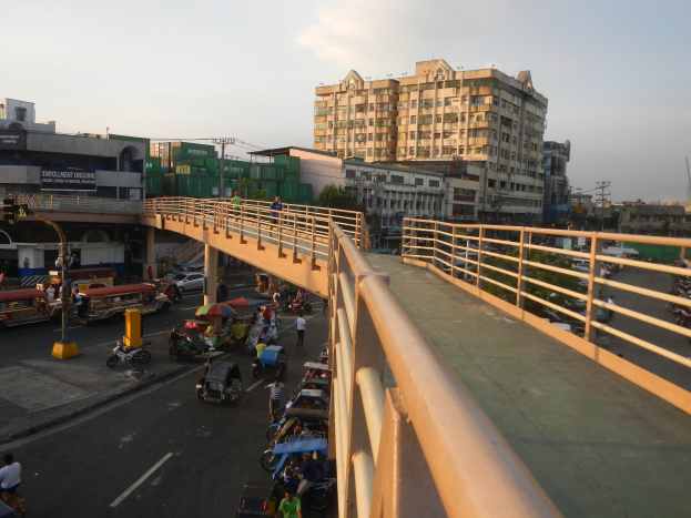 Eine Brücke über eine belebte Straße mit Fahrzeugen, Fußgängern und Schirmen, flankiert von Geländern und vor dem Hintergrund von Gebäuden, Bäumen und einem klaren Himmel.