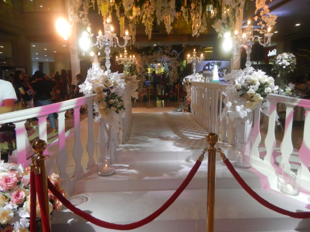 Eine Hochzeitszeremonie in einem Einkaufszentrum mit weißem Teppich, Geländern, Blumenschmuck, Kronleuchtern und Deckenbeleuchtung, bei der viele Menschen anwesend sind, einige tragen Taschen.