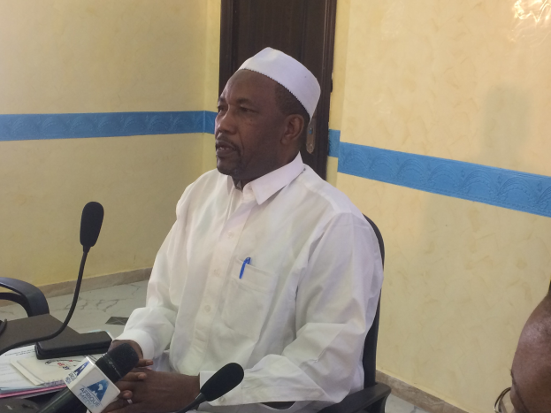 Ein Mann in einem weißen Kleid und einer Mütze sitzt an einem Tisch mit Mikrofonen und spricht, während Papiere und Gegenstände auf dem Tisch zu sehen sind, mit einer anderen Person mit Brille rechts von ihm, in einem Raum mit einer Wand und einer Tür im Hintergrund.