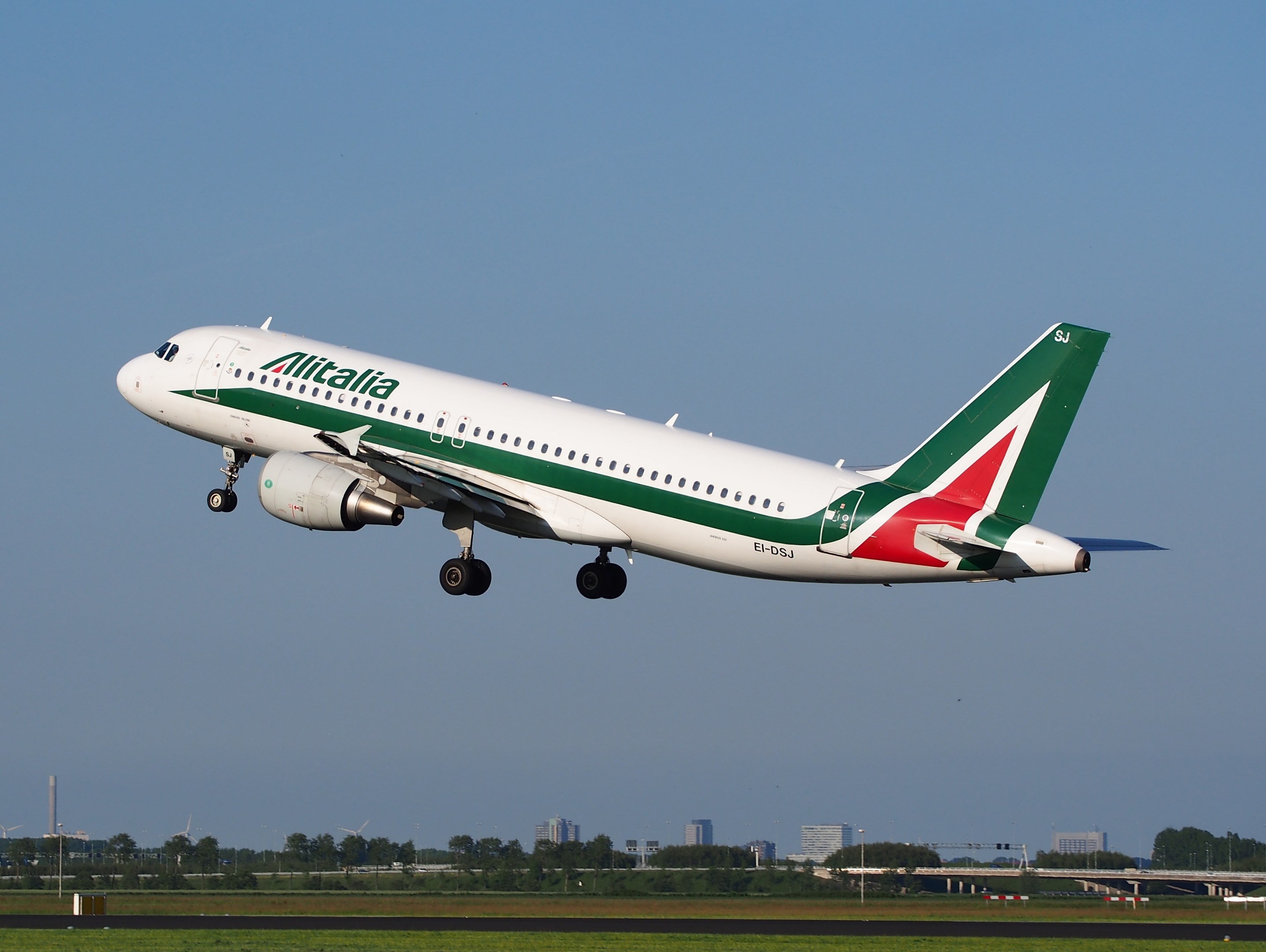 Ein Airbus A320-232 ERJ-EJZ der Alitalia, der vom Frankfurter Flughafen abhebt, mit grünem Gras, B├Ąumen, Geb├Ąuden und einem klaren blauen Himmel im Hintergrund.