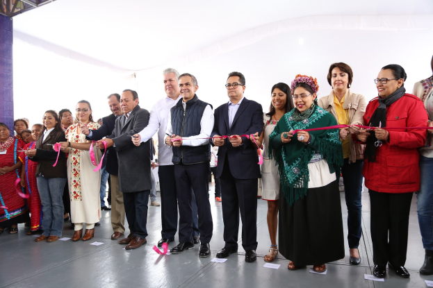 Eine Gruppe von Menschen hält eine Schere bei einer Eröffnungszeremonie, mit einer Säule links, verstreuten Papieren auf dem Boden und einem weißen Tuch im Hintergrund.