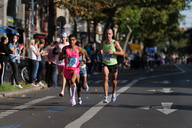 Gruppe von Läufern bei einem Marathon auf einer Stadtstraße mit Zuschauern auf der linken Seite, Bäumen, Gebäuden und einem Fahrrad im Hintergrund.