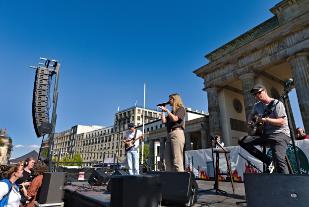 Eine Gruppe von Menschen, die auf einer Bühne vor dem Brandenburger Tor in Berlin Musik spielen, mit Lautsprechern und Equipment drumherum, vor einem klaren blauen Himmel.