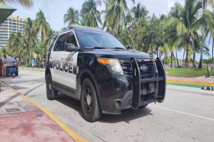 Ein Polizeiwagen am Straßenrand geparkt, umgeben von Menschen, Fahrrädern, Gras, Bäumen, Gebäuden und einem klaren blauen Himmel, mit einem sichtbaren Vorderrad.