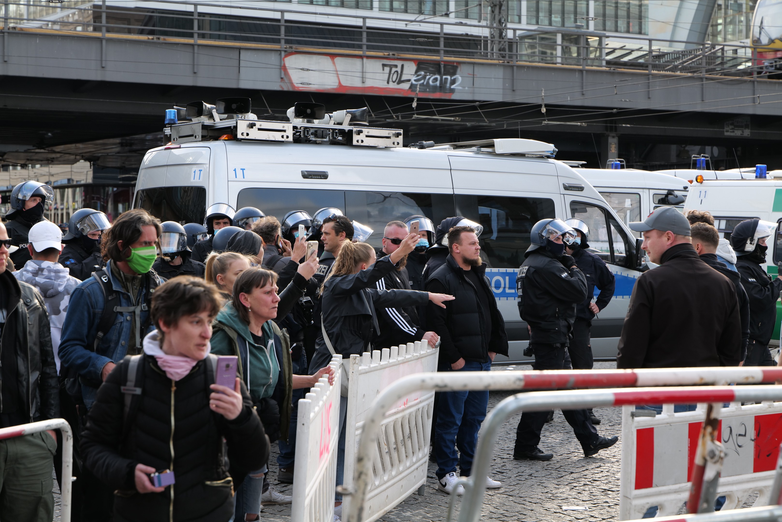Eine Gruppe von Menschen steht vor Polizeifahrzeugen, einige tragen Helme und halten Telefone, mit Barrieren im Vordergrund und einer Brücke und Gebäuden im Hintergrund während einer Protestauflösung in Berlin.