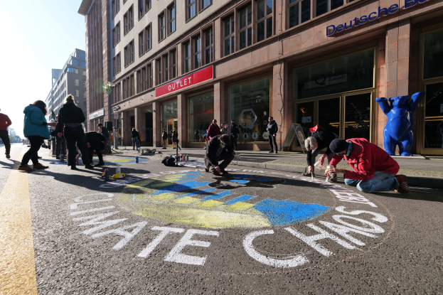 Menschen sitzen vor einem Gebäude mit Fenstern und Namensschildern, umgeben von Flaschen und anderen Gegenständen, während sie an einer Klimaprotestaktion in Berlin teilnehmen, bei der es einen klaren blauen Himmel gibt.