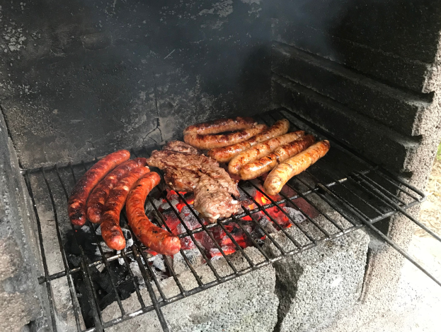 Grill mit Würstchen und Fleisch, das darauf gegrillt wird, umgeben von Kohle und Feuer, auf Gras im Freien