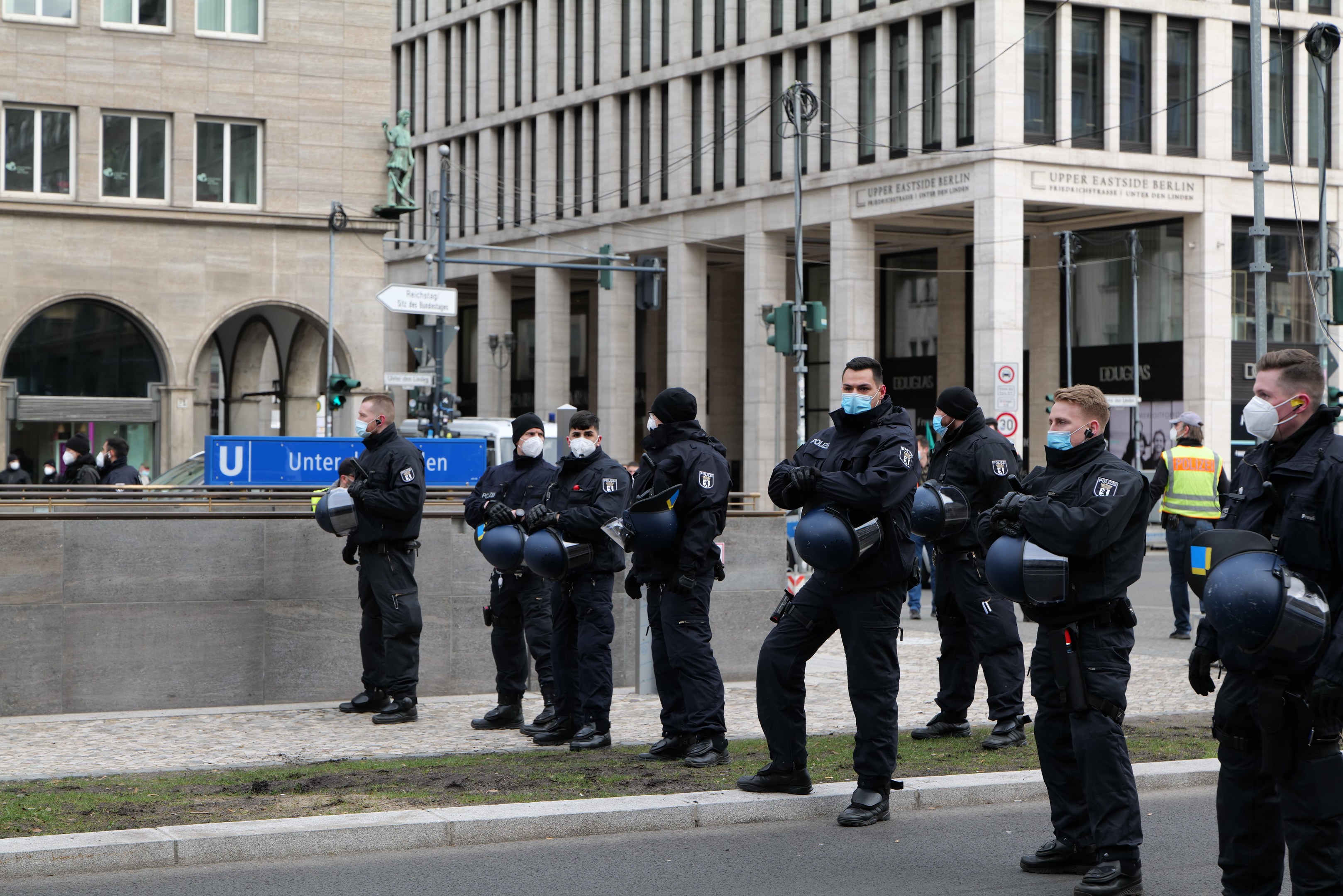 Polizeibeamte in schwarzen Uniformen und Masken stehen vor einem Berliner Gebäude mit Glasfenstern und Säulen, einige halten Helme, mit Laternen, Verkehrsampeln, Texttafeln, einer Statue und Gras im Hintergrund.