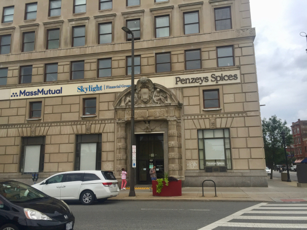 Großes Gebäude mit "Massachusetts Mutual Financial Group"-Schriftzug, parkende Autos davor, Straßenlaternen, Fahrzeuge, Fußgänger, Bäume und bewölkter Himmel.