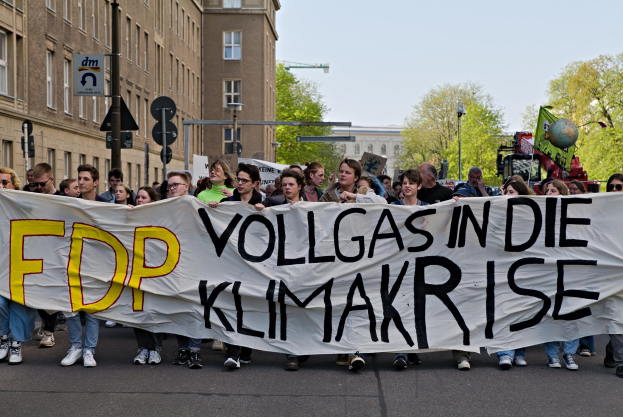 Gruppe von Menschen, die eine Straße entlanggehen und ein Banner mit der Aufschrift "FDP Vollgas in die Klimakrise" halten, mit Gebäuden, Bäumen und Verkehrszeichen im Hintergrund unter einem klaren blauen Himmel.