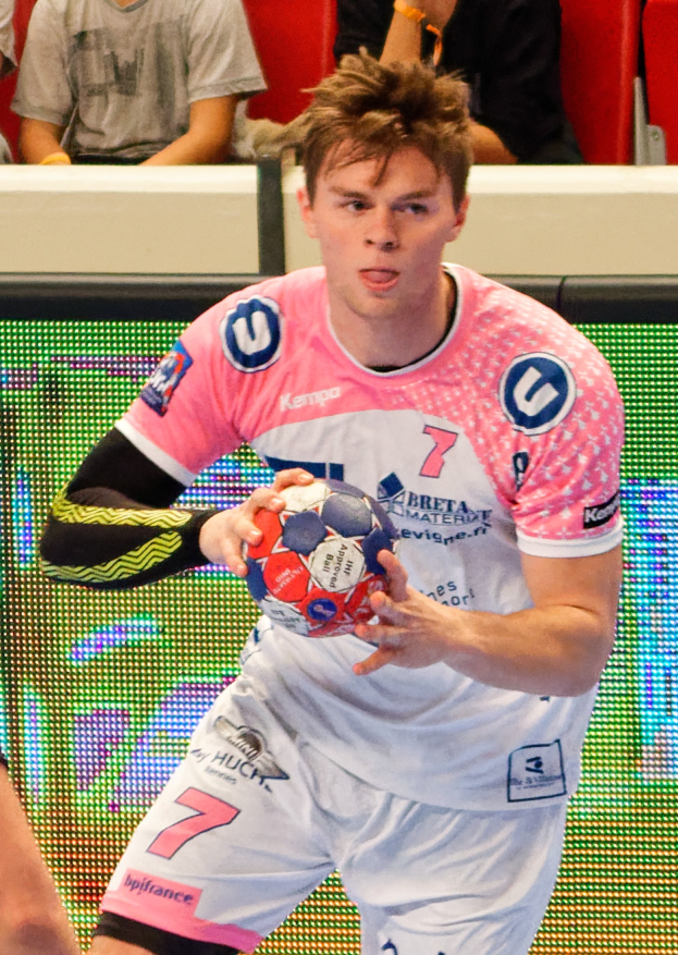 Ein Mann in einem pinken Hemd hält einen Fußball, während im Hintergrund einige Menschen auf Stühlen sitzen.
