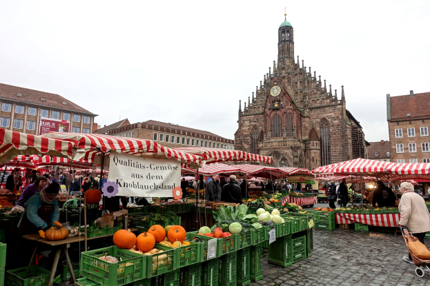 Ein belebter Markt in Nürnberg, Deutschland, mit Früchten, Gemüse, Menschen, Zelten, Gebäuden, einem Uhrenturm und Himmel.