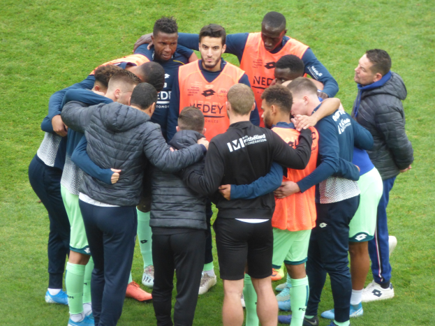 Eine Gruppe von Männern in Jacken, die im Kreis auf einem Fußballfeld stehen, wahrscheinlich Teil eines Teams.
