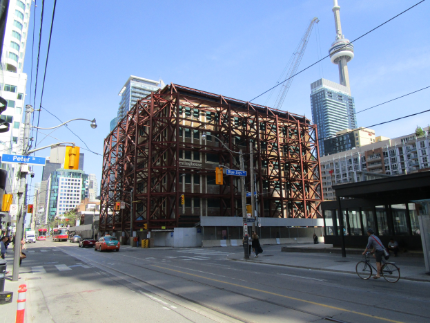 Eine belebte Stadtstraße mit einem großen Gebäude im Hintergrund, Fahrzeugen auf der Straße, einem Radfahrer, Fußgängern auf dem Gehweg, Ampeln, Masten mit angebrachten Schildern, einem Kran und dem Himmel.