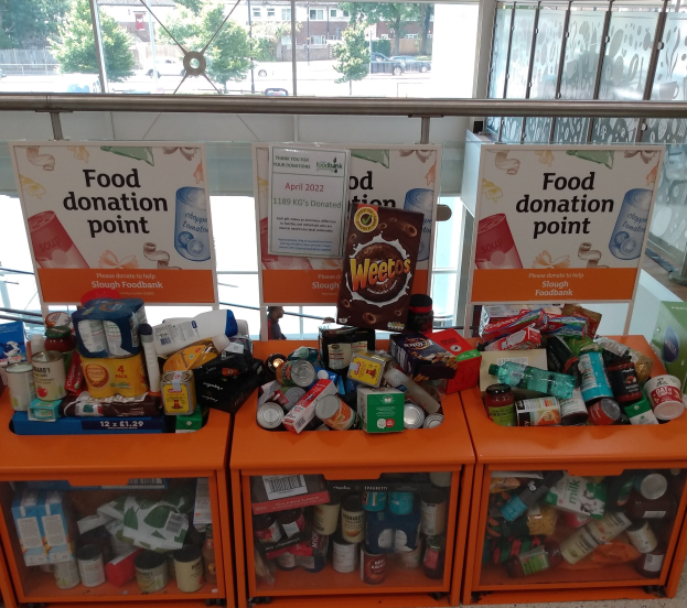 Orangene Schübe mit Lebensmittelspenden wie Flaschen, Dosen und Kisten, mit Informationsschildern und einem Blick durch Glasfenster auf Fahrzeuge, Bäume, Gebäude und Himmel.