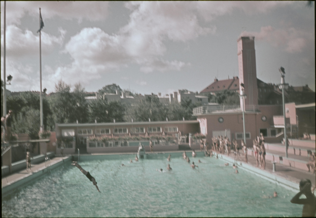 Altes Schwarz-Weiß-Foto von Menschen, die in einem Pool schwimmen, mit Gebäuden, Bäumen, Polen, Flaggen und einem Uhrenturm im Hintergrund unter einem bewölkten Himmel.