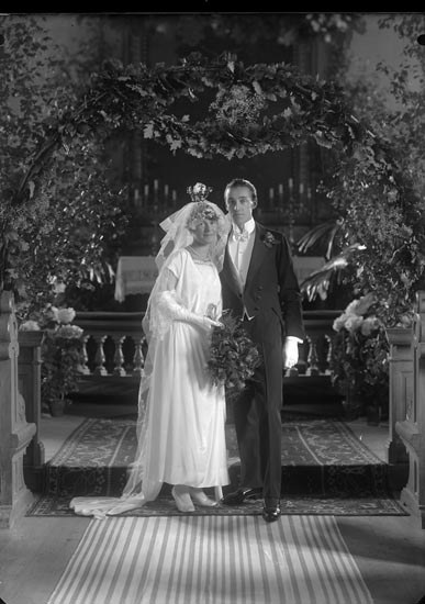 Schwarz-weiß-Foto eines Mannes und einer Frau, die unter einem Blumenschmuckbogen stehen, die Frau hält einen Strauß Blumen.