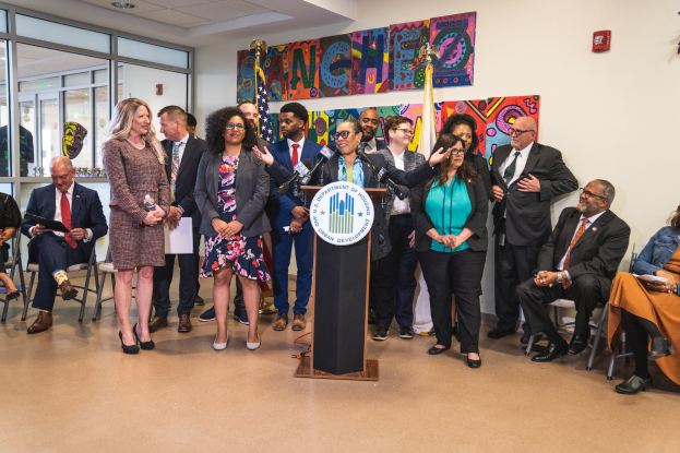 Gruppe von Menschen vor einem Rednerpult mit Mikrofon und einer Tafel mit der Aufschrift "Sancho Stadtverordneten joins City Council", mit sitzenden und stehenden Teilnehmern, Flaggen, Feuerlöscher und Deckenleuchten im Hintergrund.