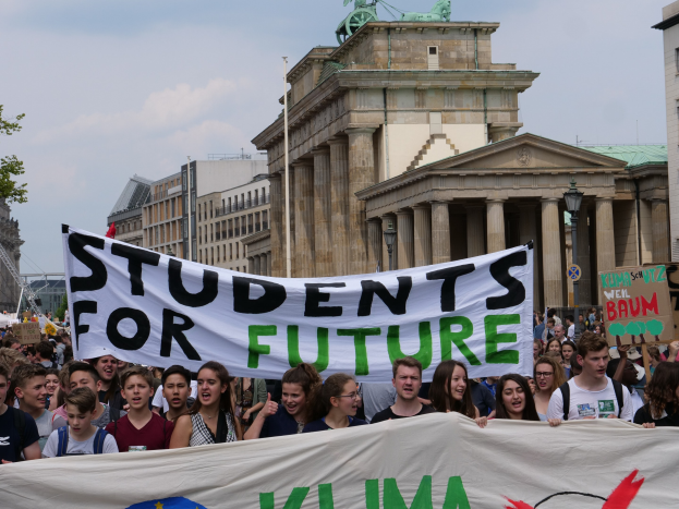 Eine Gruppe von Studenten marschiert in Berlin, hält eine bunt bemalte "Students for Future"-Schleife gegen den Hintergrund von Gebäuden, Bäumen und Himmel.