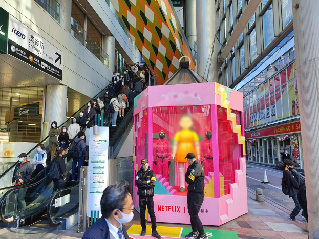 Eine Gruppe von Menschen steht vor einem gläsernen Gebäude mit Säulen und Texttafeln, einige tragen Masken, während eine Rolltreppe mit Menschen zu sehen ist; ein pinker Kasten in der Mitte zeigt Schaufensterpuppen und trägt die Aufschrift "Netflix's new Netflix booth in Tokyo."