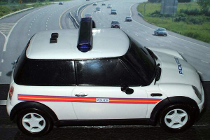 Ein weißer Polizeiwagen steht im Vordergrund mit mehreren Fahrzeugen, die sich dahinter auf der Straße bewegen, umgeben von Gras und Bäumen auf beiden Seiten.