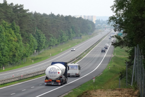 Ein großer Lkw fährt auf einer von beiden Seiten von Bäumen gesäumten Autobahn, mit einem Zaun und Gras auf der rechten Seite und im Hintergrund sichtbaren Gebäuden unter einem klaren blauen Himmel.