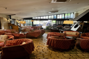 Ein geräumiger Eingangsbereich des Hotel de Ville mit zahlreichen Stühlen und Tischen, gepolsterten Sofas, Wandlampen, einer Deckenklimaanlage und großen Glasfenstern an der Rückseite.