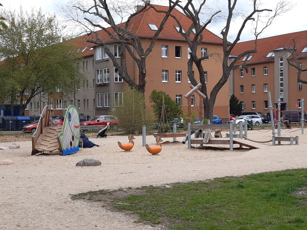 Ein Spielplatz mit einer Rutsche, Schaukeln und anderen Geräten, umgeben von Gras, Bäumen, Gebäuden, Fahrzeugen auf einer Straße und einem bewölkten Himmel.