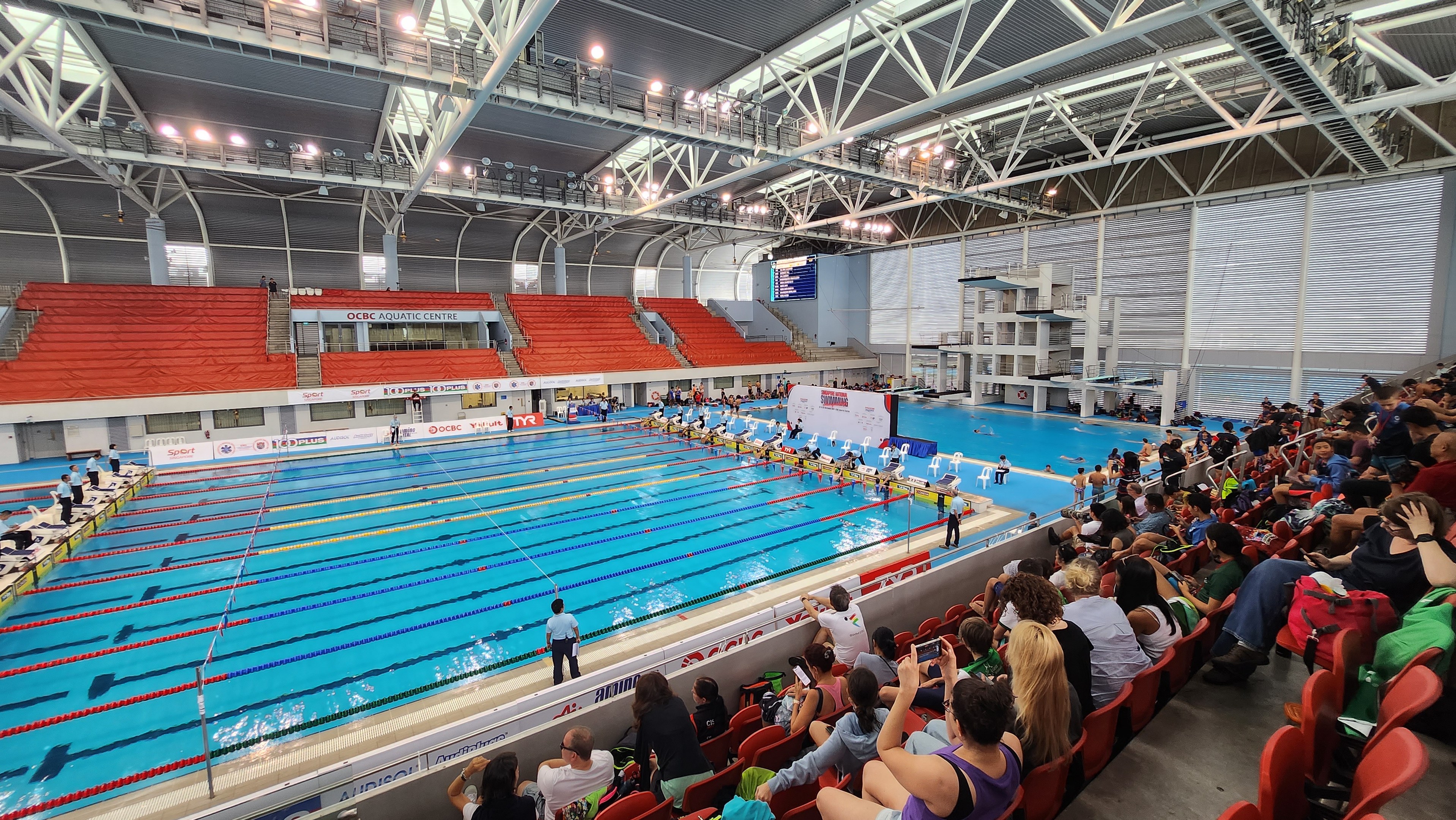 Großes Hallenbad mit vielen Menschen auf Stühlen und einigen im Wasser Stehenden, mit hoher Decke mit Lampen und Eisenstangen und Hintergrundtafeln mit Text, was auf das Olympische Schwimmzentrum in London hinweist.