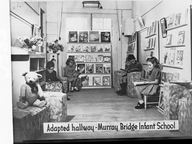 Eine Gruppe von Kindern sitzt auf Stühlen in einem Raum mit Bücherregalen, einem Teddybär links, einer Blumenvase auf dem Tisch und gerahmten Gegenständen an der Wand, mit dem Text "Angepasster Flur Murray Bridge Kindergarten" unten.