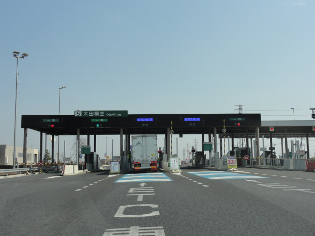 Eine Mautstelle mit einem vor ihr parkenden Lastwagen, neben einer mit wei├č-blauen Linien markierten Stra├če, Lichtmasten, Schildern und anderen Gegenst├Ąnden, mit Geb├Ąuden, B├Ąumen und einem klaren Himmel im Hintergrund.