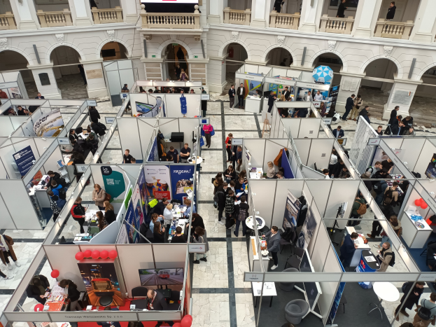 Große Gruppe von Menschen auf einer internationalen Messe mit Ständen, Bannern und Luftballons in einem Raum mit gewölbter Architektur.
