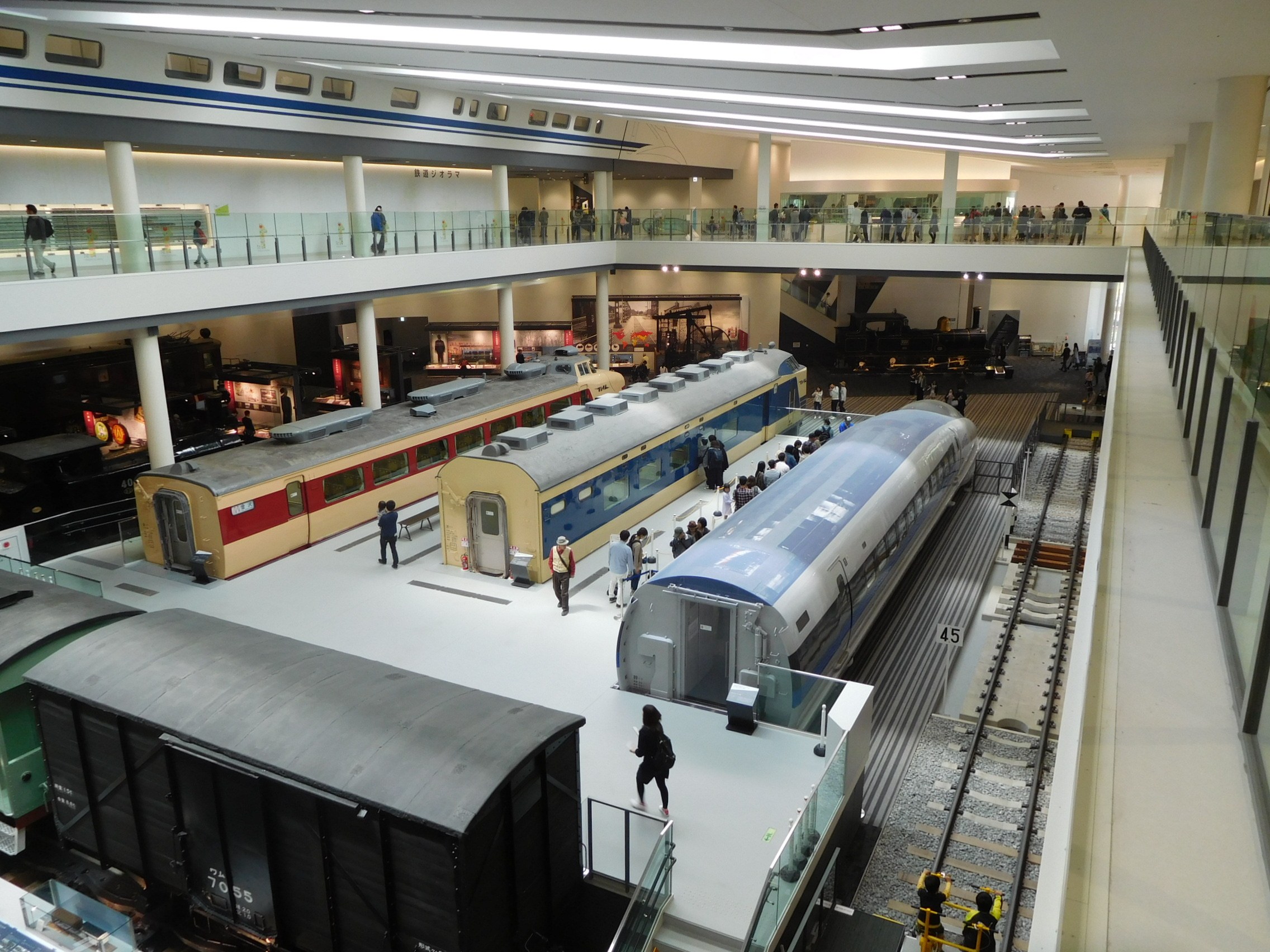 Eine großzügige Museumsausstellung mit mehreren Zügen in verschiedenen Größen und Farben, die von Besuchern vom Boden, Geländern und Säulen unter Deckenbeleuchtung betrachtet werden.