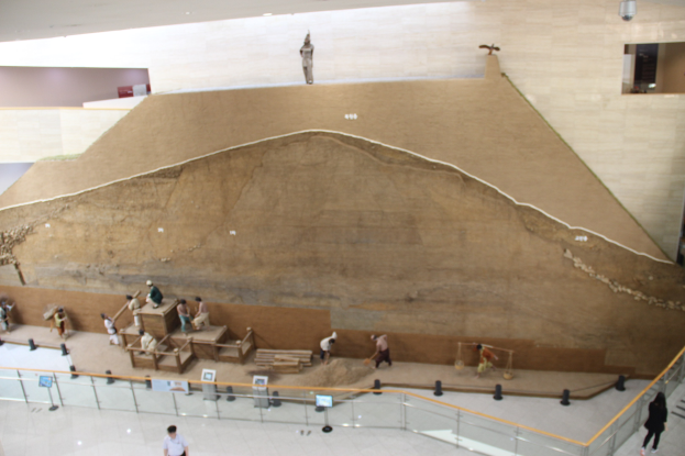 Große Felsformation als Grabhügel in einer Museumsausstellung umgeben von Informationsschildern und Besuchern hinter einem Zaun.