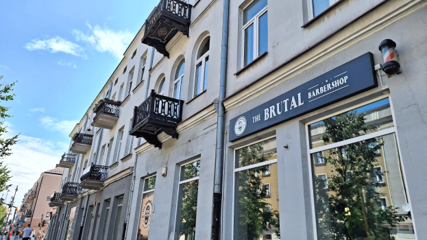 Brutalistischer Barbershop-Laden in Berlin mit Betongebäuden, Fenstern, Geländern, Balkonen und Namensschildern, umgeben von Bäumen, Laternenmasten und Fußgängern unter einem bewölkten Himmel.
