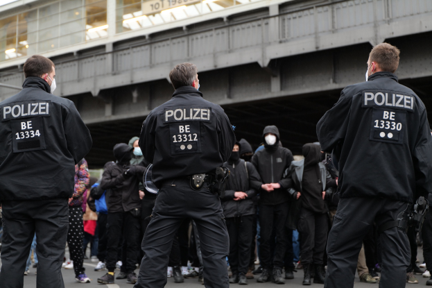Polizeibeamte in schwarzen Uniformen und Masken vor einer Menge während einer Demonstration, mit einer Brücke und einem Gebäude im Hintergrund.