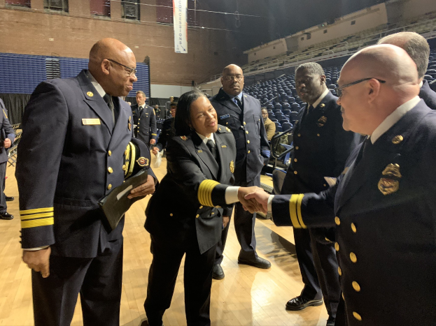 Gruppe uniformierter Personen, die sich die Hände schütteln, vor sitzenden und stehenden Menge, mit einem Banner im Hintergrund, das "Feuerwehrchef und Offizier des Jahres" verlässt.
