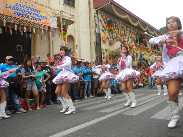 Eine Gruppe von Frauen in pinken und weißen Outfits tanzt auf der Straße, umgeben von einer Menge Schaulustiger, mit Gebäuden, Bäumen und einem klaren blauen Himmel im Hintergrund.