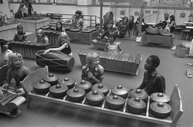 Schwarz-weißes Bild von Kindern, die Musikinstrumente in einem Raum mit Stühlen, Tischen, Pflanzen, gerahmten Wänden und Deckenleuchten spielen.